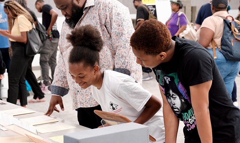 Three people lean over a table, looking at papers and smiling, while others in the background walk around and talk in a bright, busy indoor setting.