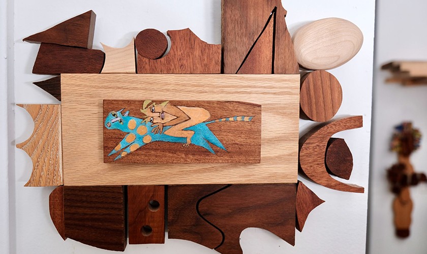 A wooden art piece features varied geometric shapes around a central panel showing a stylized figure riding a blue, spotted creature. The wood is of different shades and textures, creating a layered, abstract design.