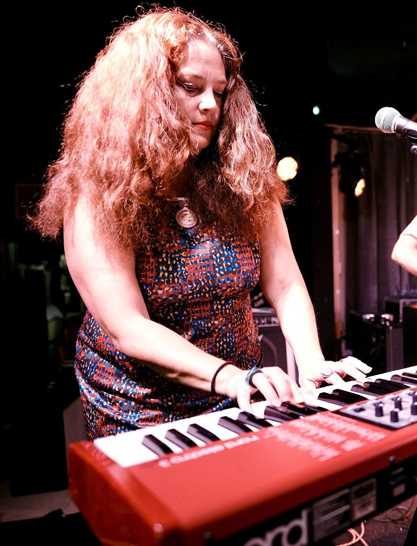 A woman with long, curly hair wearing a patterned dress plays a red keyboard on stage, focused on the keys, with stage lights in the background.