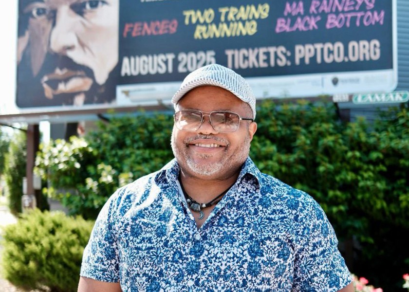 A smiling man in a blue patterned shirt holds a coffee cup, standing outside in front of a large billboard advertising August Wilson’s American Century Cycle Experience, listing plays and ticket information for August 2025.