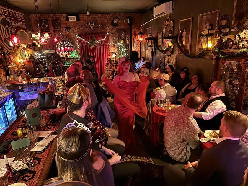 A crowd of folks sit in a bar watching a burlesque dancer in a red dress and scarf walking in the middle of the room