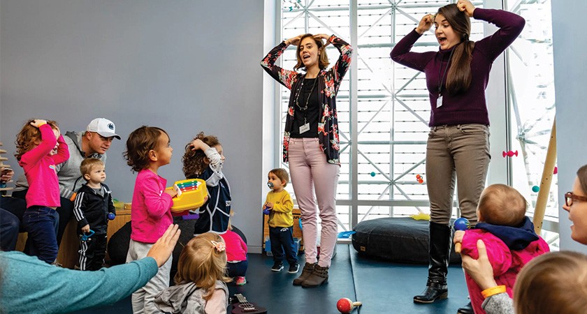 Two adults stand beside each other with their hands raised up to their head in front of a crowd of children